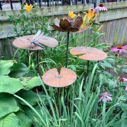 Metal lily pads rise through flowers and green plants. A metal dragonfly sits on one lily pad. A metal lily is beside them.